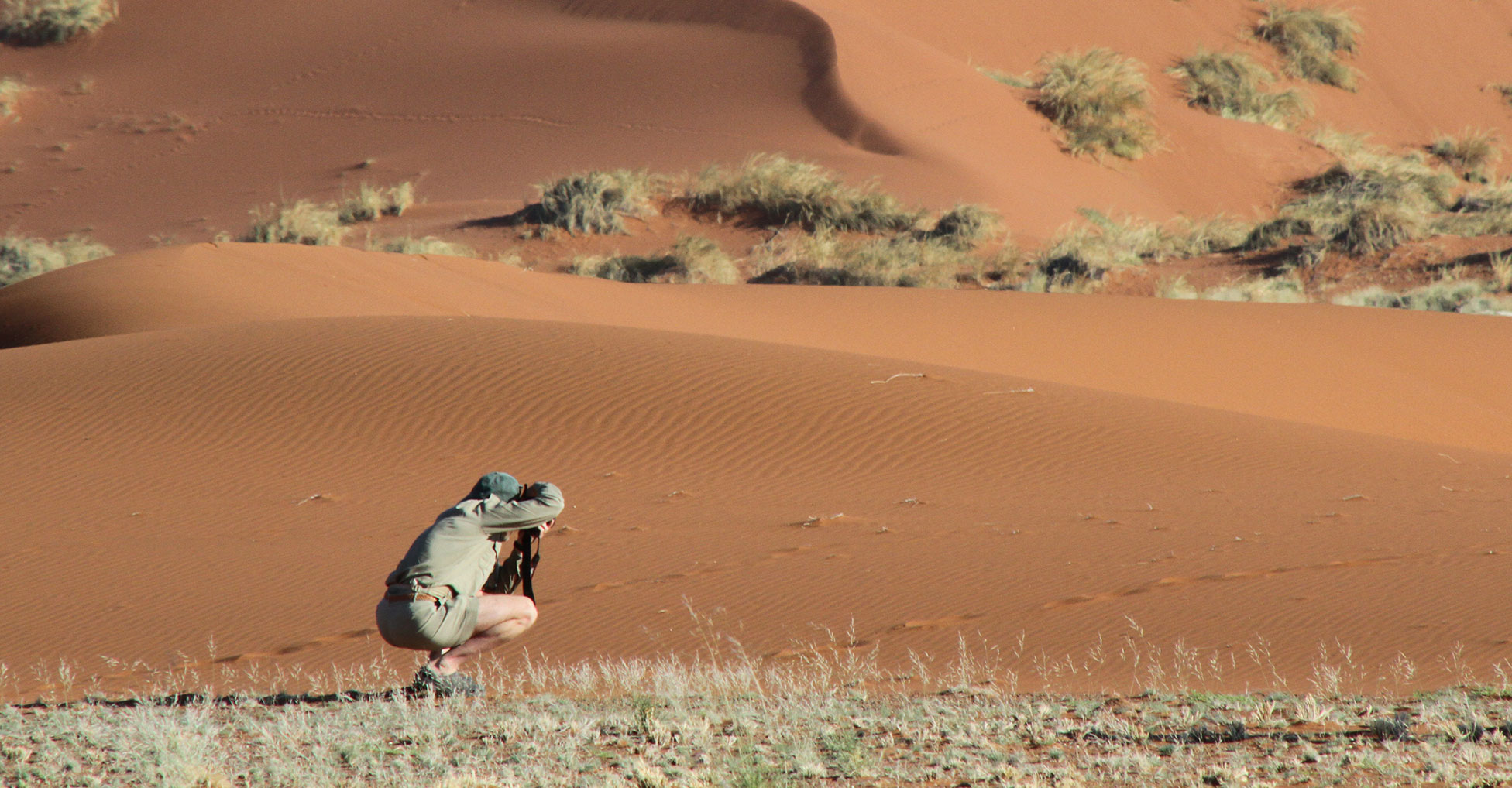  A person crouches near the base of a sand dune while aiming a camera offscreen