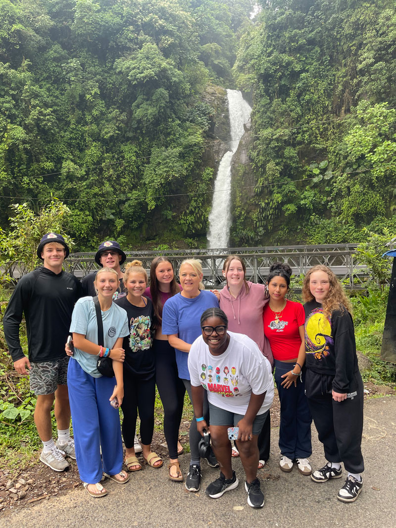 A group of nine teenagers and one adult smile and pose in front of a waterfall