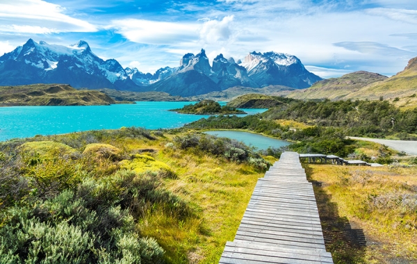 A wooden walkway leads through a valley landscape with a lake and snow-capped mountains.