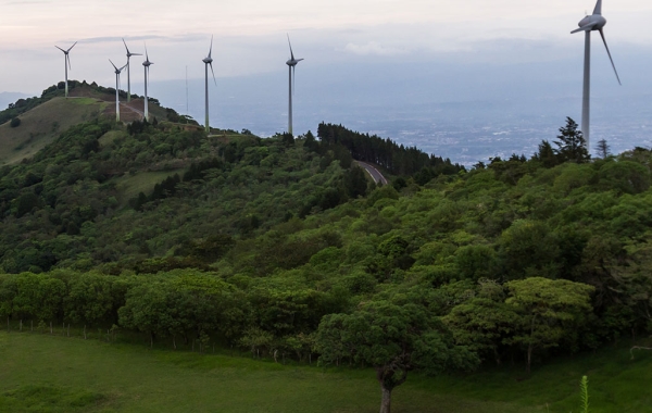 A series of seven wind turbines sit atop the ridge of a tree-covered hill, with a valley and clouds in the distance