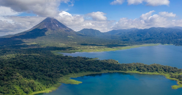 Aerial view of a lake surrounded by forest, with a volcano and low mountains in the background