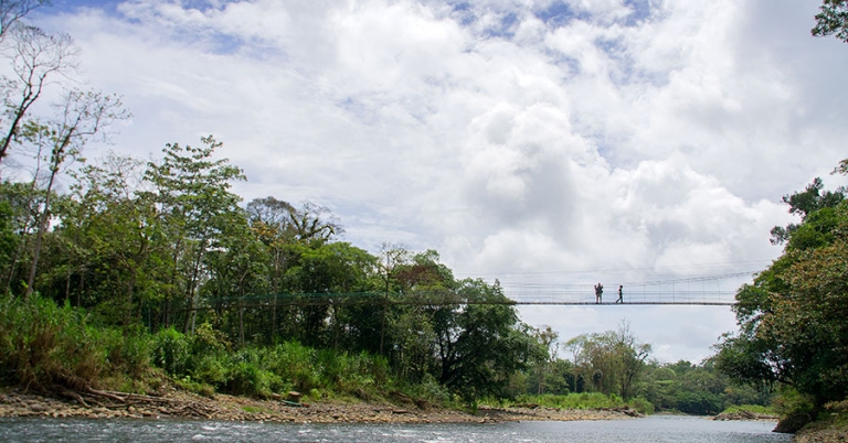 Two people in silhouette cross a suspension bridge over a river