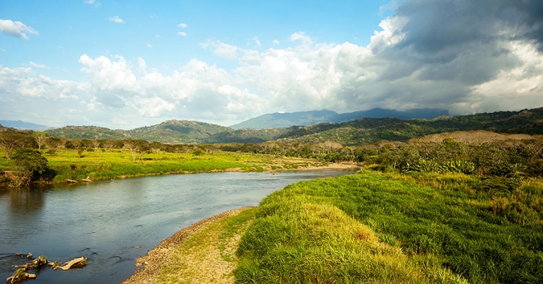 Panoramic view of a wide river with vegetation on the banks and low mountains in background