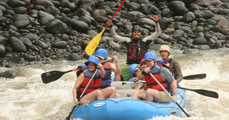 Four passengers and a guide hold oars and smile while sitting in an inflatable raft on a river with small waves