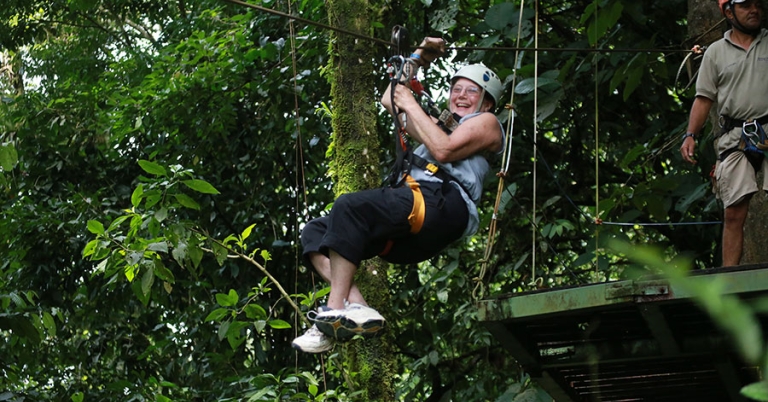 A person wearing a harness hangs suspended from a cable with a metal platform and trees in the background