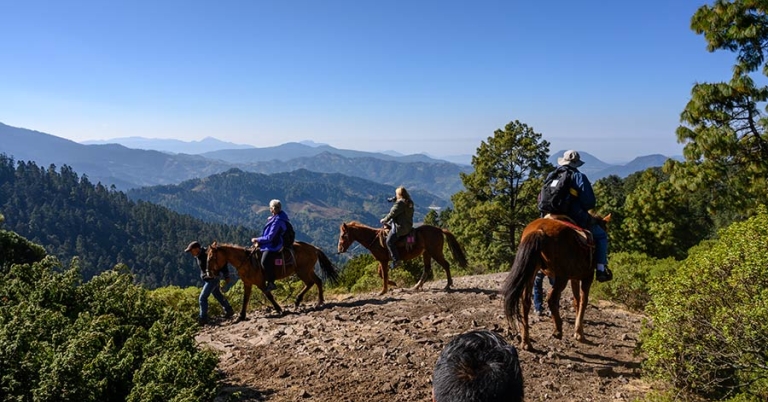 Three people ride horseback near an overlook with a mountainous landscape in the background