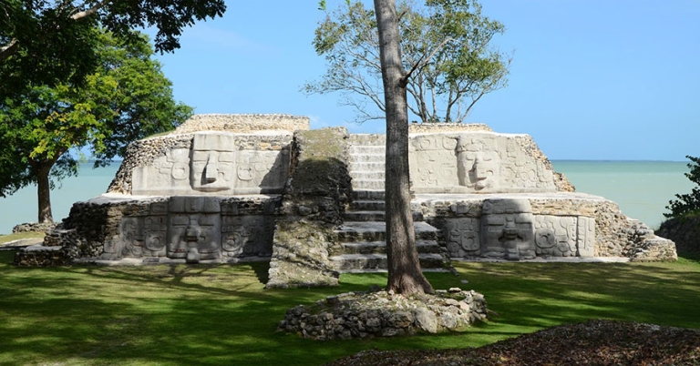 Two-tiered Maya pyramid with four large masks flanking a stairway and the ocean in the background