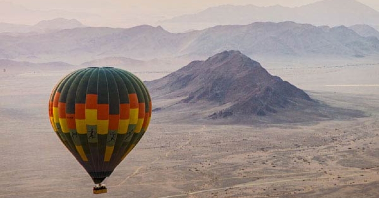 A hot air balloon floats in the sky over a desert landscape with low mountains in the background