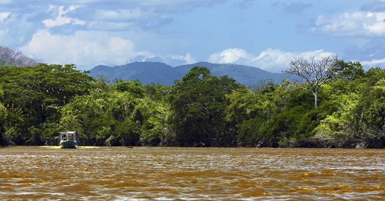 A small boat on a wide river, with trees and low mountains in the background