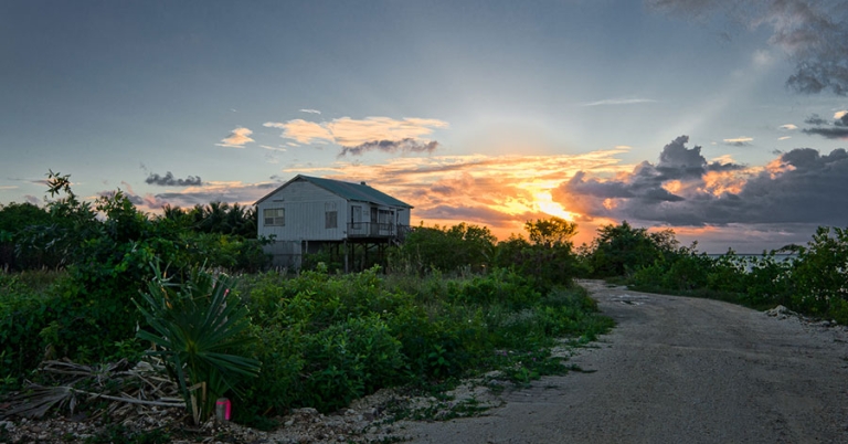 The sun sets over a sandy road surrounded by vegetation and a house on stilts in the background