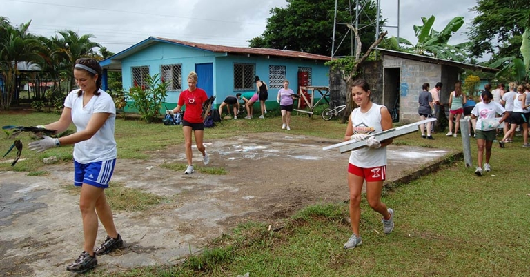 Several teenagers wearing gloves carry paint supplies and building materials with a school building in the background