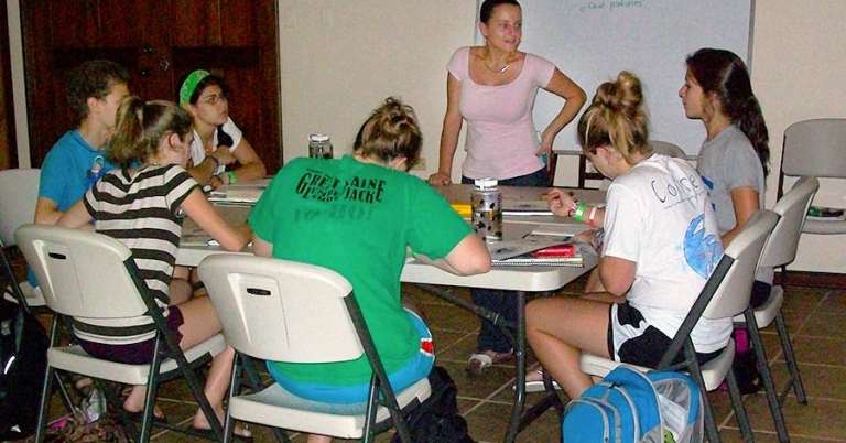 A teacher stands in front of a dry-erase board with Spanish text written on it, while six teenage students sit around a table