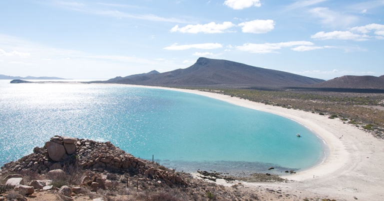 Landscape of the wide, circular bay with light blue water, a sandy shoreline, and a low mountain in the background
