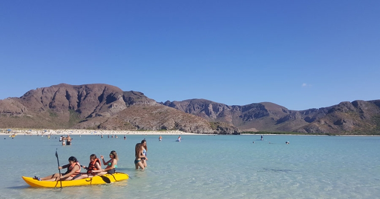People wade and kayak in a shallow bay with hills in the background