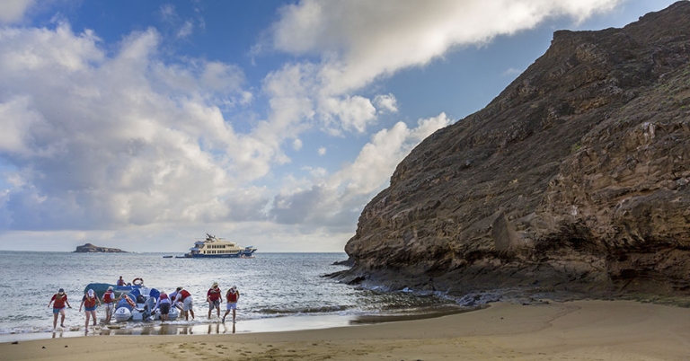 Eight or nine people wearing life jackets walk in ankle-deep water away from a panga resting on a sandy beach, with a larger boat in the ocean behind them