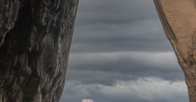 A tall rock formation rises out of the ocean in the distance, framed by an opening in a closer rock formation