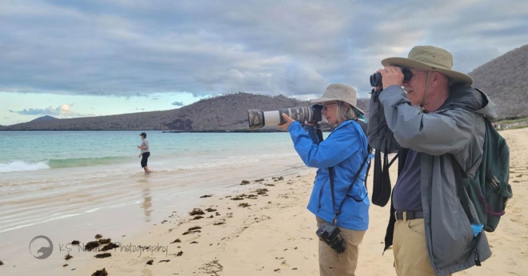 Two people stand side-by-side on the beach, one looking through binoculars and the other through a camera