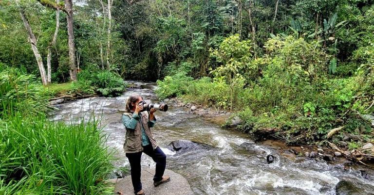 Krisztina Scheeff aims her camera offscreen while standing near a river in the forest
