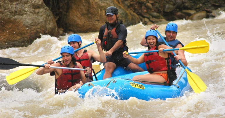 Four people and a guide hold oars while sitting in an inflatable raft on a river with small waves