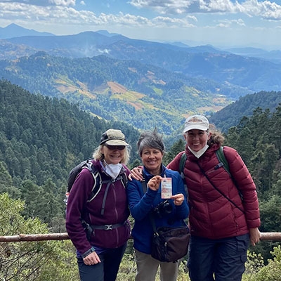 Three people stand smiling in front of a wooden handrail overlooking a mountain landscape in the background