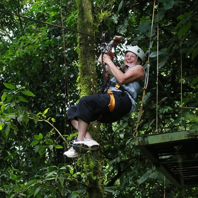 A person wearing a harness hangs suspended from a cable with a metal platform and trees in the background