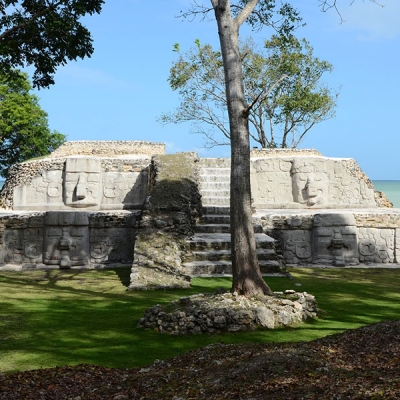 Two-tiered Maya pyramid with four large masks flanking a stairway and the ocean in the background