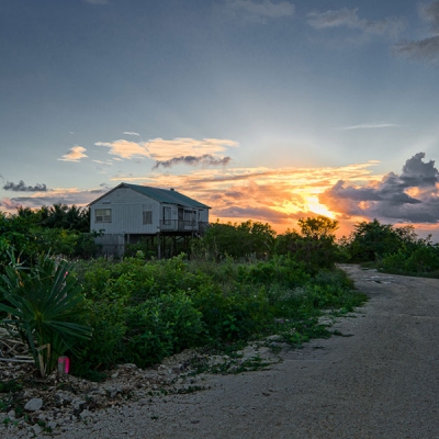 The sun sets over a sandy road surrounded by vegetation and a house on stilts in the background