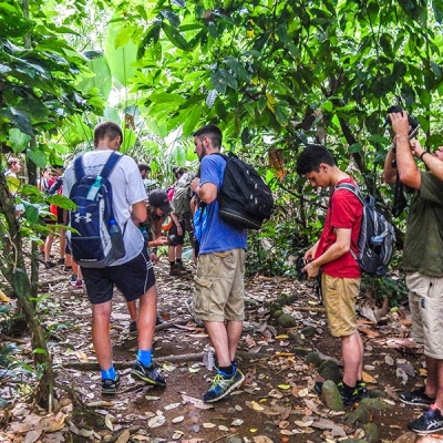 A group of teenage students walk and take pictures on an unpaved rainforest trail