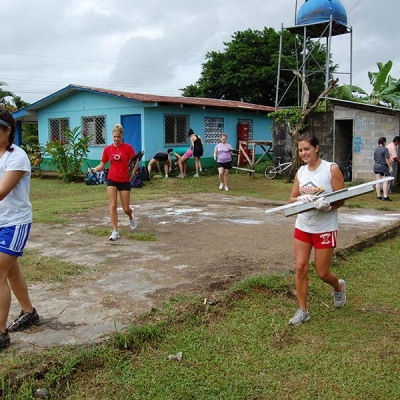 Several teenagers wearing gloves carry paint supplies and building materials with a school building in the background