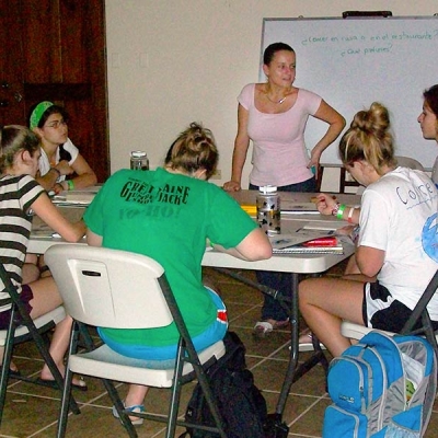 A teacher stands in front of a dry-erase board with Spanish text written on it, while six teenage students sit around a table