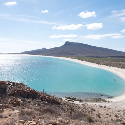 Landscape of the wide, circular bay with light blue water, a sandy shoreline, and a low mountain in the background