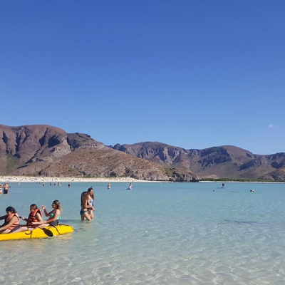 People wade and kayak in a shallow bay with hills in the background