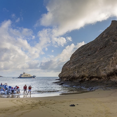Eight or nine people wearing life jackets walk in ankle-deep water away from a panga resting on a sandy beach, with a larger boat in the ocean behind them