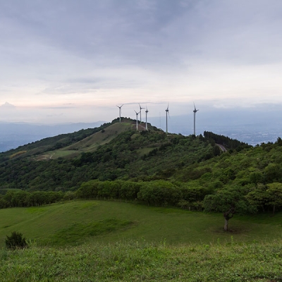 A series of seven wind turbines sit atop the ridge of a tree-covered hill, with a valley and clouds in the distance