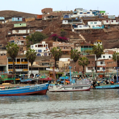 Several small, colorful fishing boats are docked near a shore line, with buildings on a hill in the background