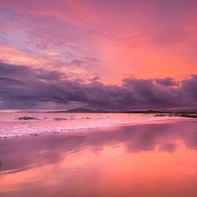 A sandy beach with pink sky and clouds reflected in the water below