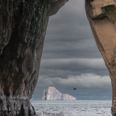 A tall rock formation rises out of the ocean in the distance, framed by an opening in a closer rock formation