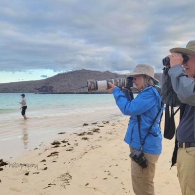 Two people stand side-by-side on the beach, one looking through binoculars and the other through a camera