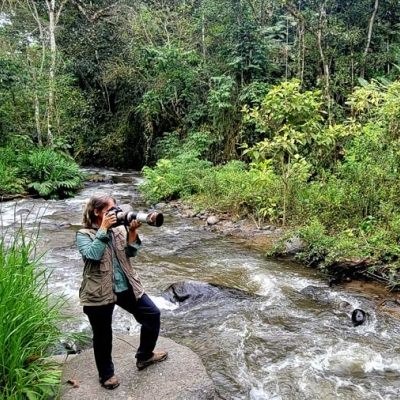 Krisztina Scheeff aims her camera offscreen while standing near a river in the forest