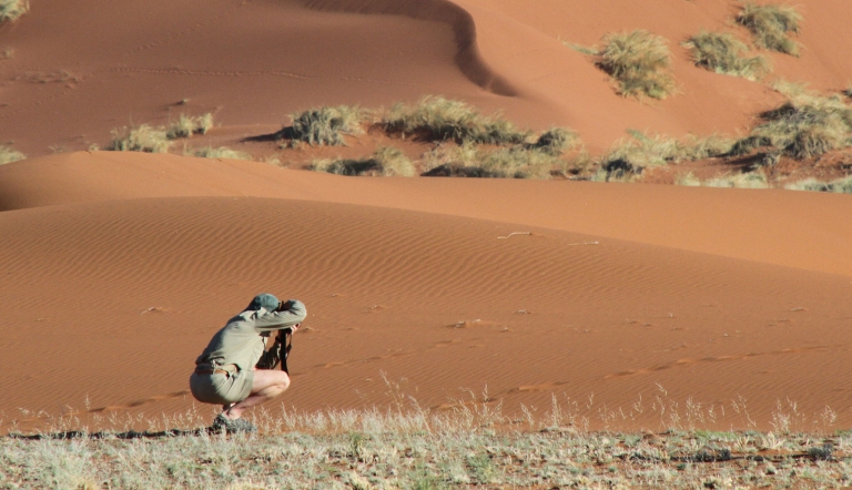  A person crouches near the base of a sand dune while aiming a camera offscreen