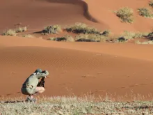 A person crouches near the base of a sand dune while aiming a camera offscreen