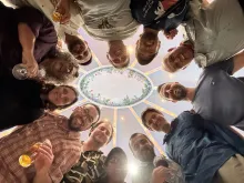 Upward view of 12 people standing in a circle, smiling down at the camera, with an ornate ceiling above them